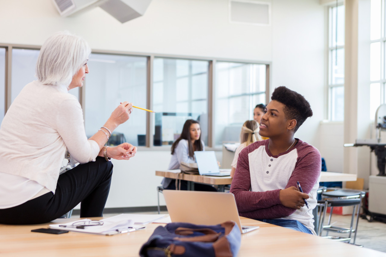 A smiling teenage boy sits at a table in his high school classroom and looks up as he listens to his senior female teacher explain the homework.
