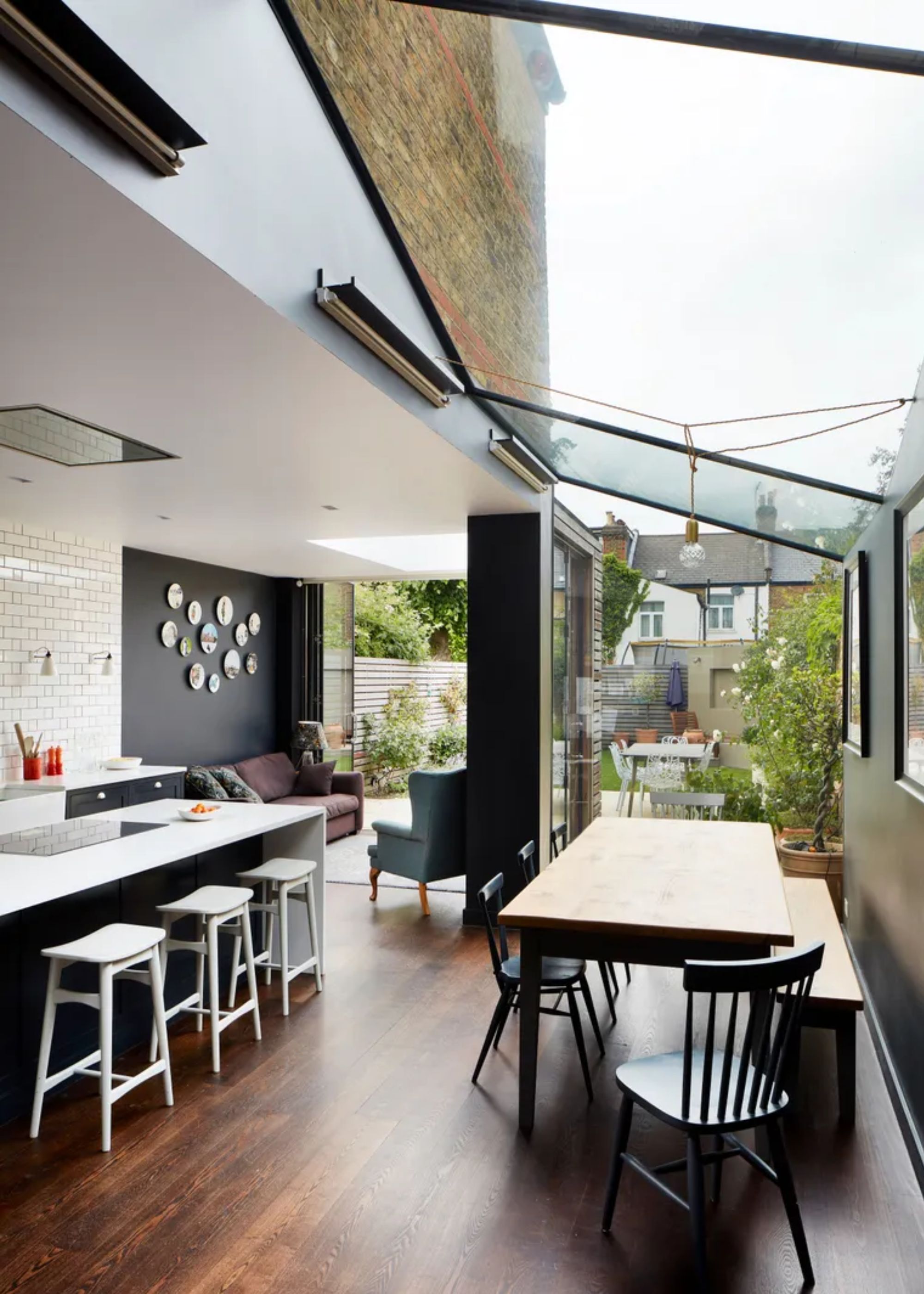 A fameless glass ceiling above a kitchen. In the kitchen is also a dining table with chairs as well as a large kitchen island with white bar stools.