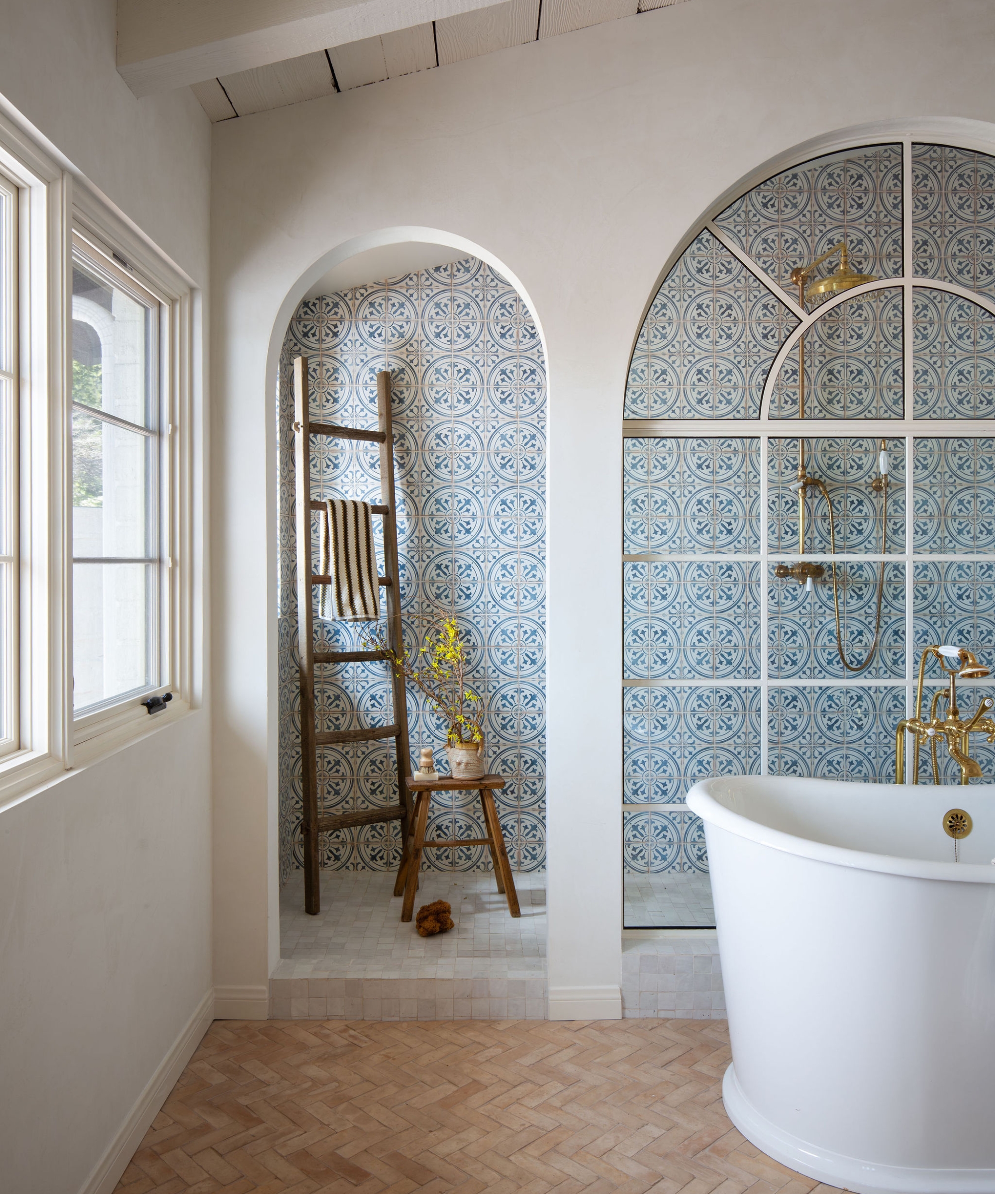 A warm white bathroom with a freestanding bath, terracotta brick floor tiles in a herringbone layout, a large shower enclosure with an arched window, and shower walls in blue and white patterned tiles
