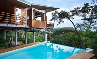 A villa in Costa Rica. Concrete columns support the house, which is covered in light wooden boards, with lots of windows and terraces. There is an infinity pool next to the villa. The villa looks over to the nature beyond.