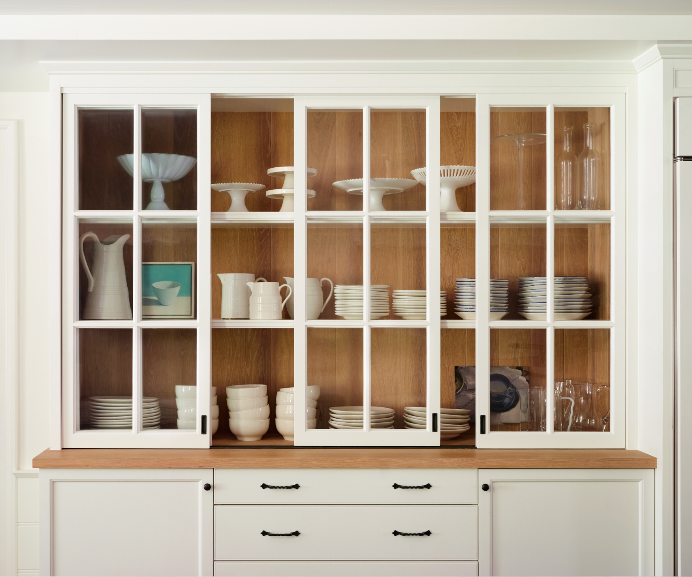 A built-in kitchen hutch painted warm white with aged bronze hardware and wooden interiors