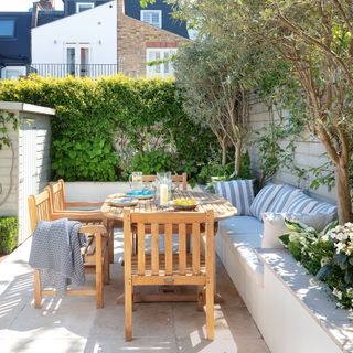 Warm wood dining table in corner of a small garden with built-in bench lined with blue and white striped cushions