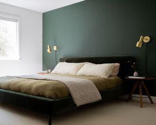 A bedroom with dark green feature wall behind dark green bed, and brass bedside wall lamps