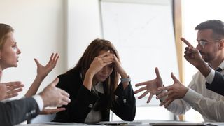 A telephoto shot of a businessperson holding their head in their hands in a boardroom, as people argue around them to represent conflict in the workplace.