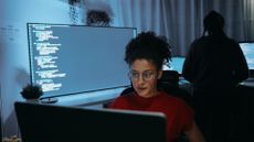 Female software developer coding on a desktop computer in a dimly-lit office space with computer code pictured on large screen in background.
