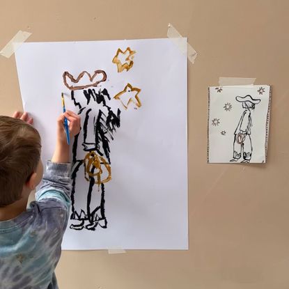 Young child painting picture of cowboy surrounded by stars, coping picture on wall beside them