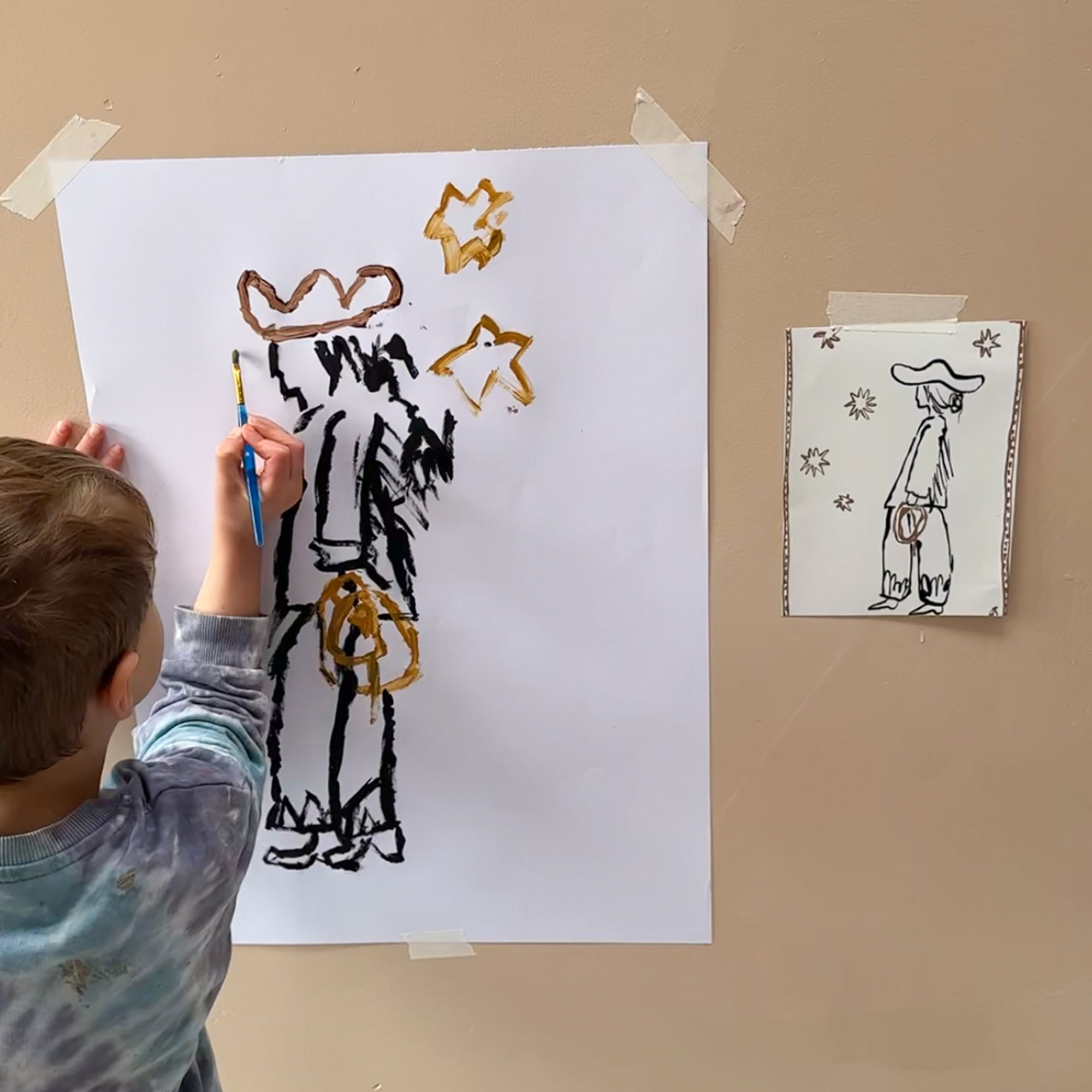 Young child painting picture of cowboy surrounded by stars, coping picture on wall beside them