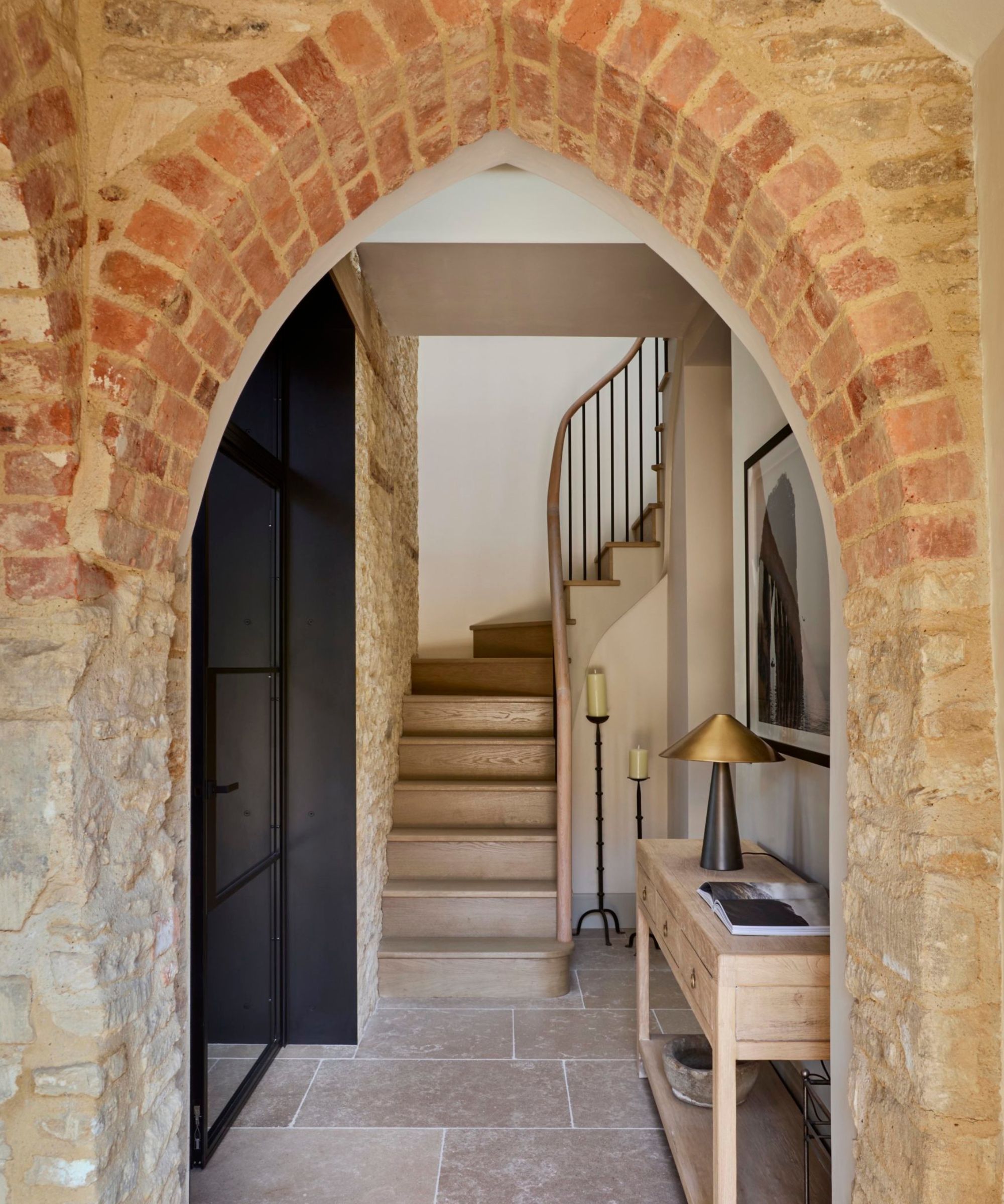 Gothic brick arch frames a black steel door opening into a hallway with oak staircase, stone floor, wooden console table and brass lamp.
