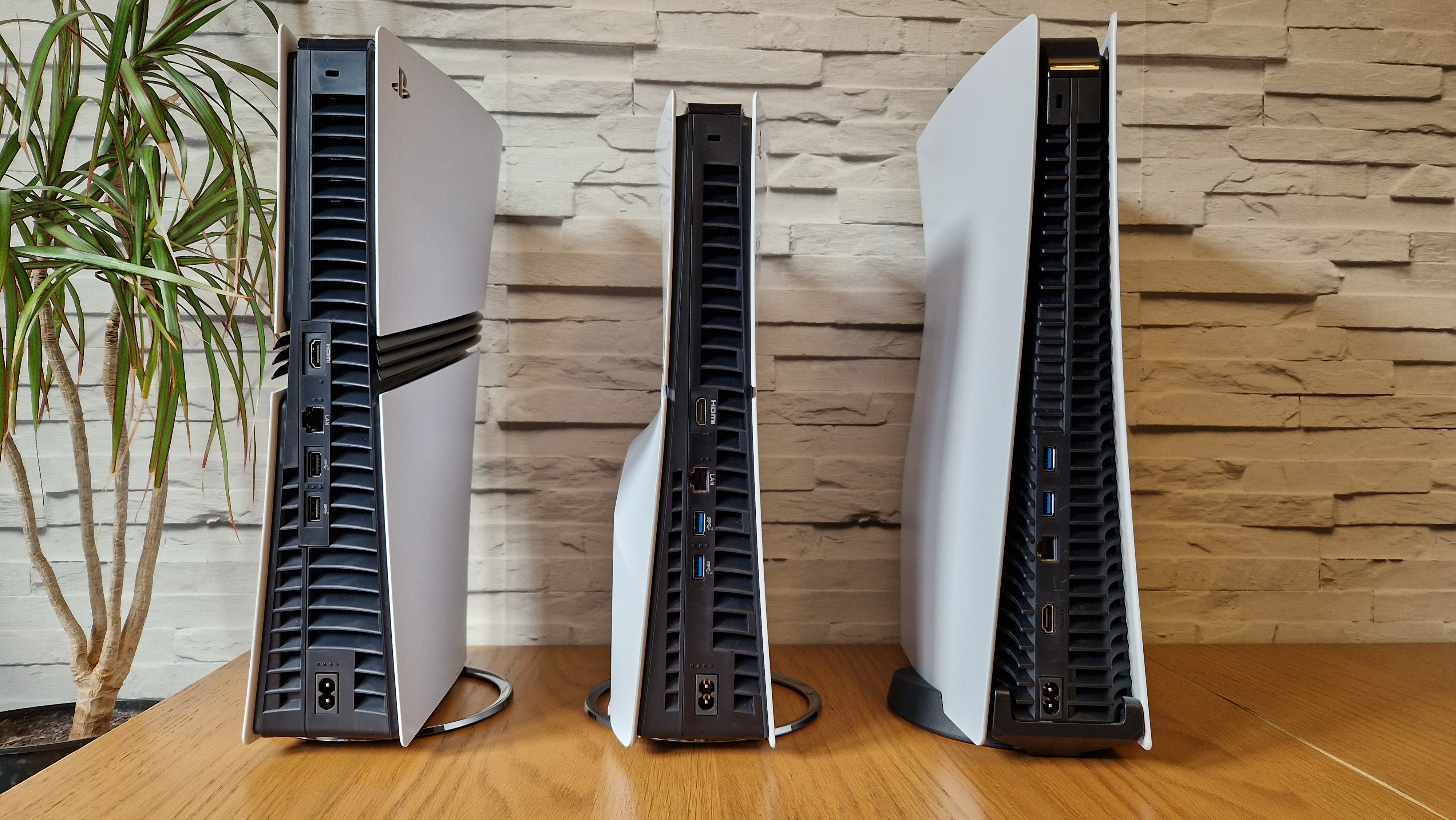 The PS5 Pro console next to a PS5 Slim and launch PS5 on a wooden desk in front of a white brick background and next to a potted plant