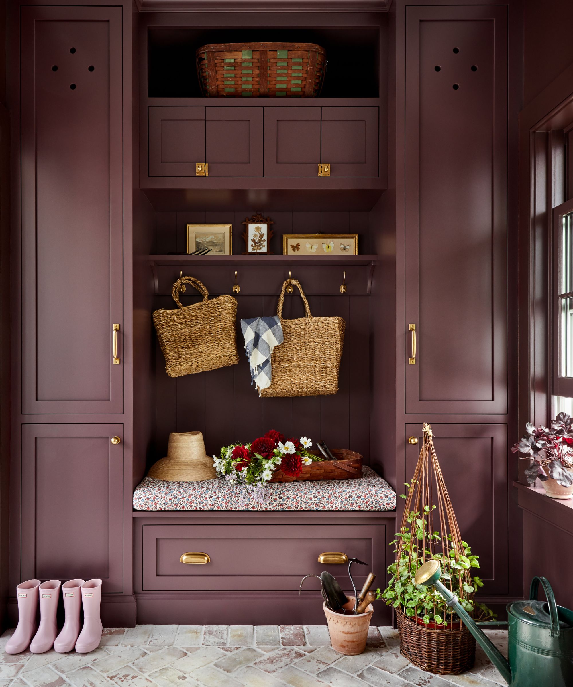A mudroom with dark purple floor-to-ceiling cabinets with brass hardware, gray brick flooring, and coat hooks in the middle of the cabinets with bags, hats, and decorative objects.
