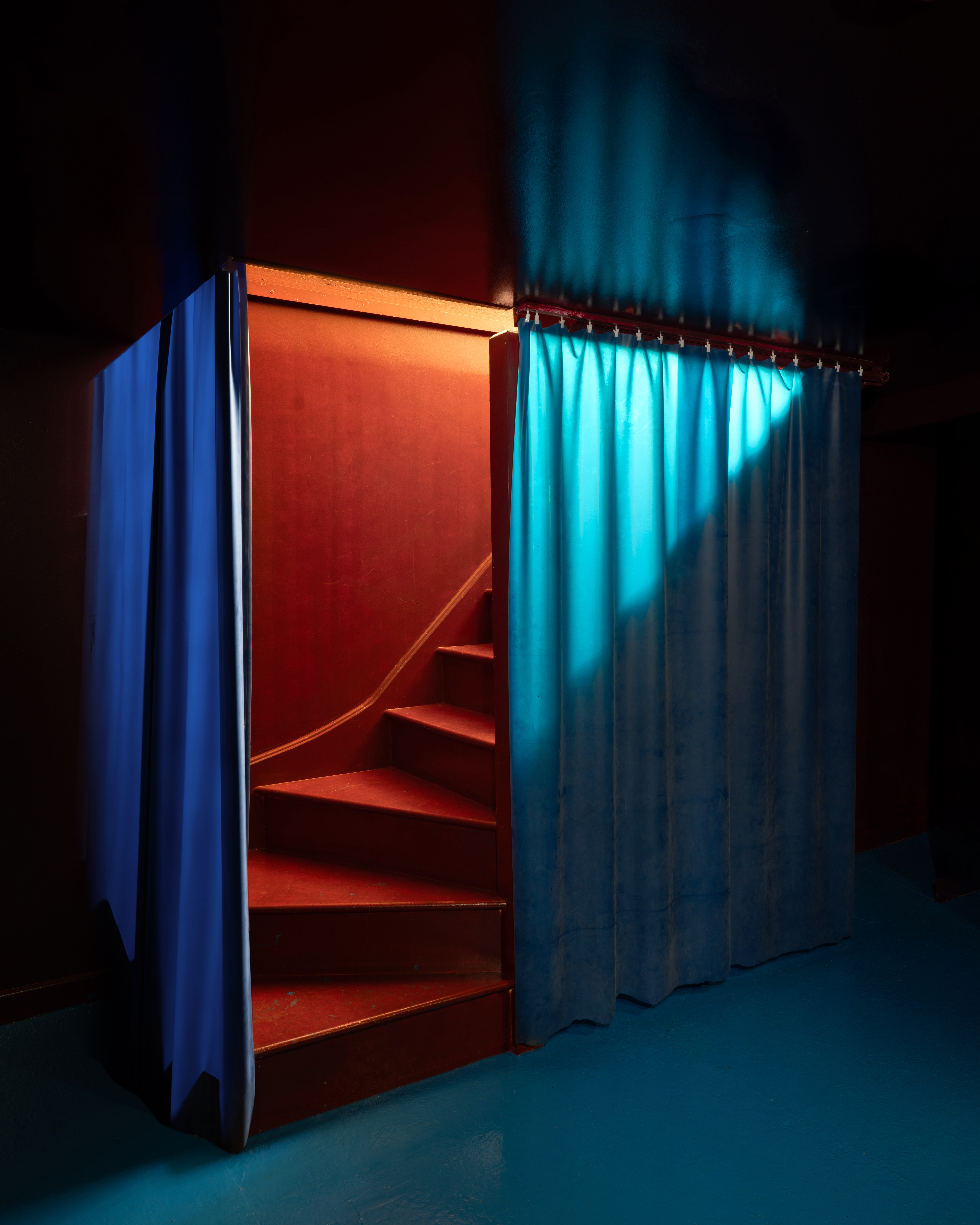 The interiors of a restaurant with red-painted floors, blue, butter yellow, and cream surfaces, figurative artwork in vibrant colors, pendant lighting with a candle-effect, and silver details.
