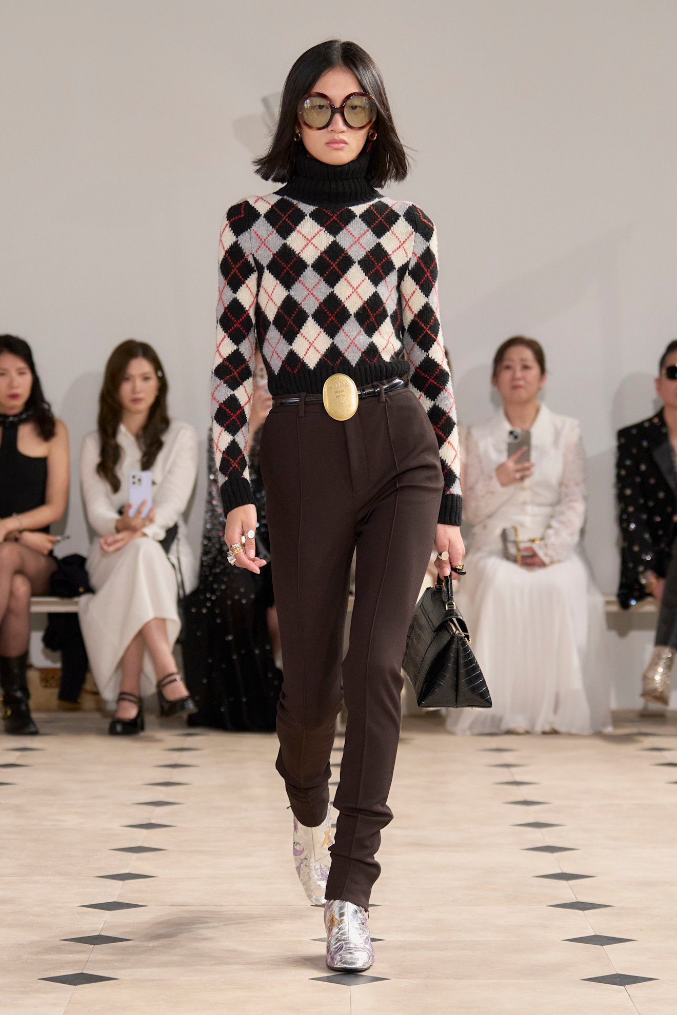 a model at Celine Spring 2026 wearing a black, white, and ivory argyle sweater and brown pants