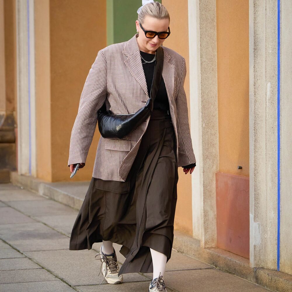 woman wears a maxi brown dress, a blazer, sneakers, and a cross-body handbag