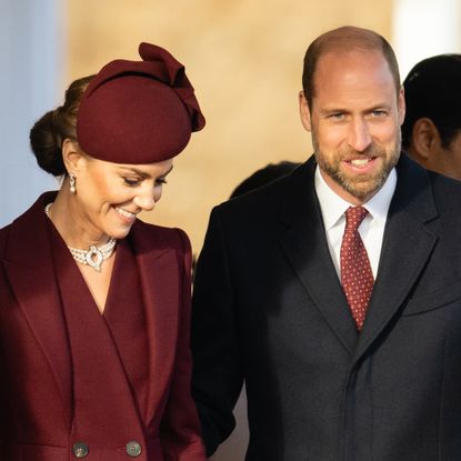 Princess Kate wearing a maroon coat and hat standing next to Prince William and smiling 