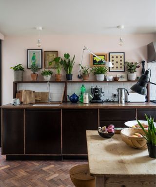 A kitchen with dark wood lower cabinets and matching open shelf above filled with potted plants and framed prints hung on the wall behind, a lighter wood table at the center, mid-tone wood parquet flooring, and subtle pink paint color on the walls