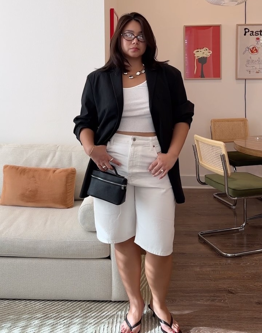 Style influencer Marina Torres poses in her living room wearing oval optical glasses, a black blazer, white tank top, white denim Bermuda shorts, mini black bag, and black kitten-heel flip-flips.