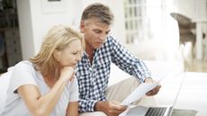 A mature couple sit in their living room, reviewing their finances on paperwork and their laptop.