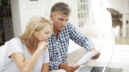 A mature couple sit in their living room, reviewing their finances on paperwork and their laptop.