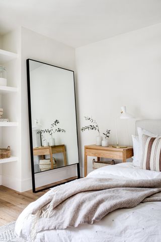 white bedroom with modern rustic side table and wooden floor, open shelving, black contemporary floor mirror, white bedding, oatmeal blanket, white table lamp