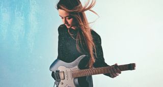 Camilla Sperati is pictured against a textured light blue background and holds her gray/blue Strandberg Boden headless guitar.