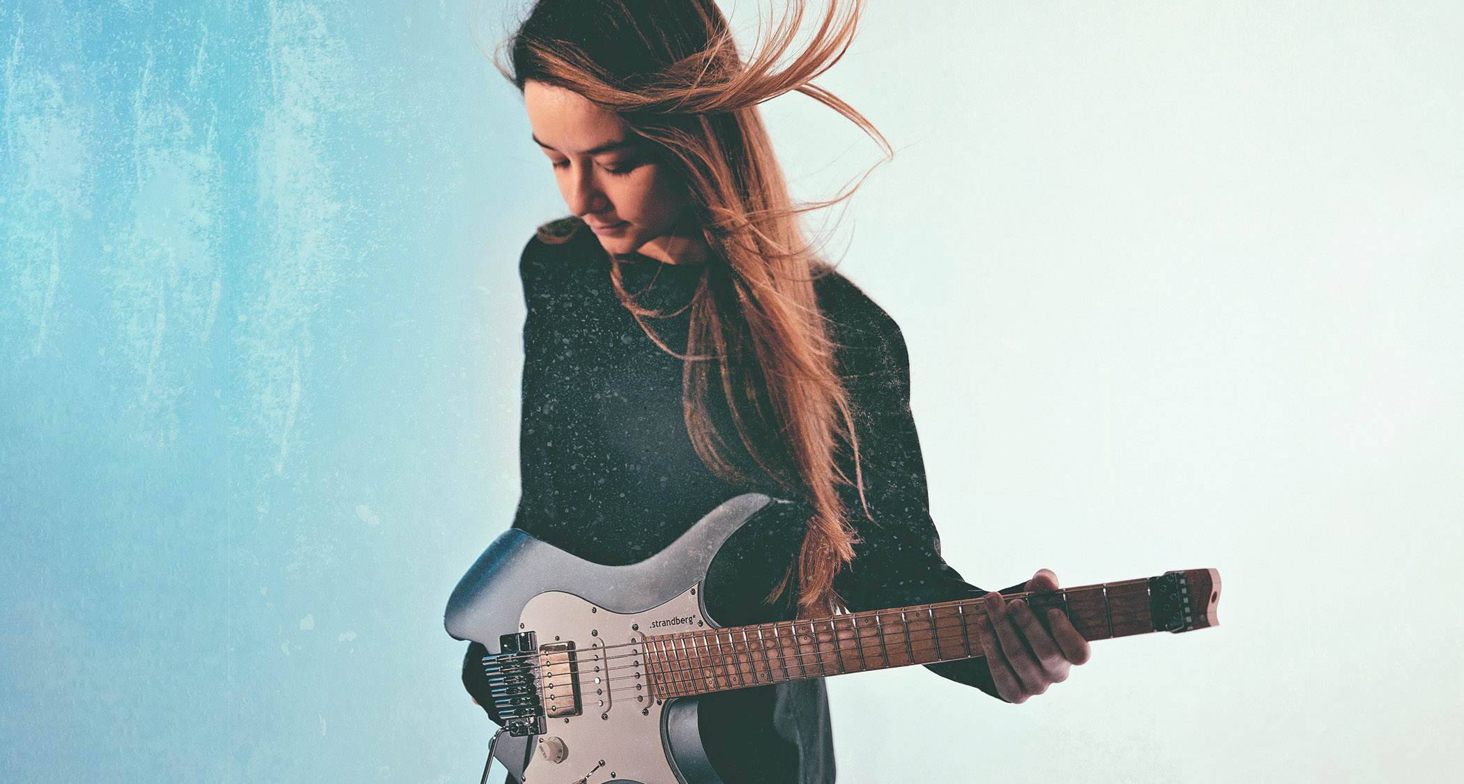Camilla Sperati is pictured against a textured light blue background and holds her gray/blue Strandberg Boden headless guitar