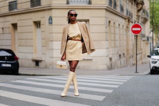 Emilie Joseph wears black large sunglasses, a pale yellow high neck / sleeveless / short dress, a camel shiny leather small belt, a beige with gold buttons blazer jacket, a beige wicker and white shiny leather bicolored handbag, pale yellow matte leather block heels / high knees boots , during a street style fashion photo session, on April 30, 2023 in Paris, France.