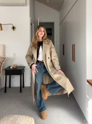 Kylie Garbourg wearing brown Ugg boots, blue jeans, a tan trench coat, a black cardigan, and a white tee shirt in a bedroom room