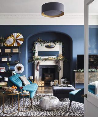 A Christmas-themed living room with blue wall paint decor, alcove with wood-burning stove, animal print rug, metallic pouffe and velvet blue sofas