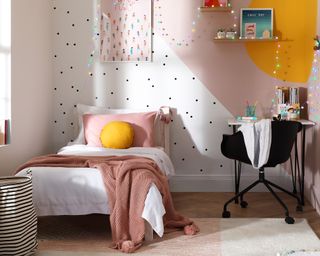 A bedroom with single bed, round yellow cushion decor, color blocked feature wall, pink throw with pom pom