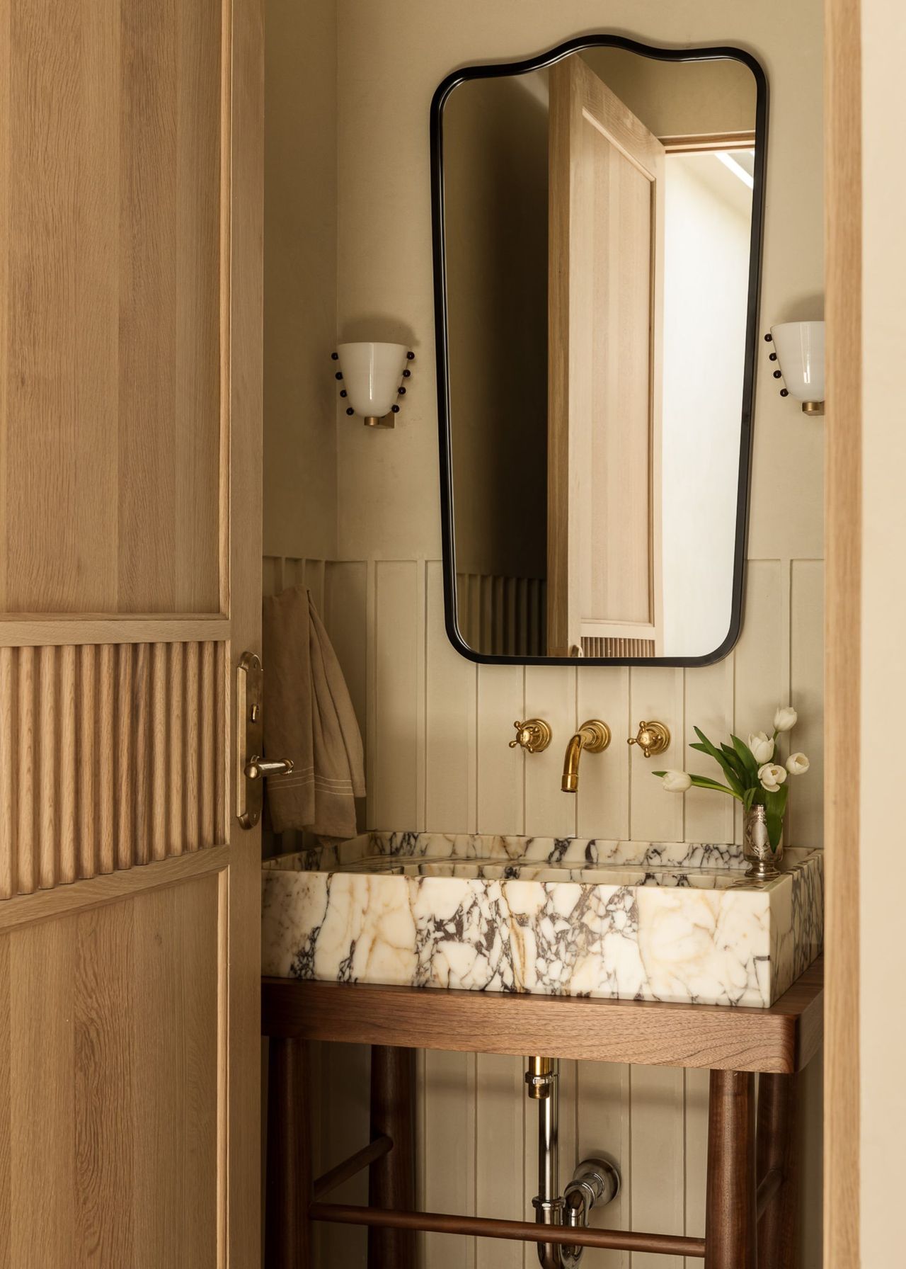 A powder room with white paneling halfway up the wall, an exposed vanity unit with a wood frame and marble sink and a mirror above it with a wall light either side