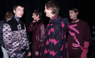 A group of male models wearing burgundy & pink clothing stand talking
