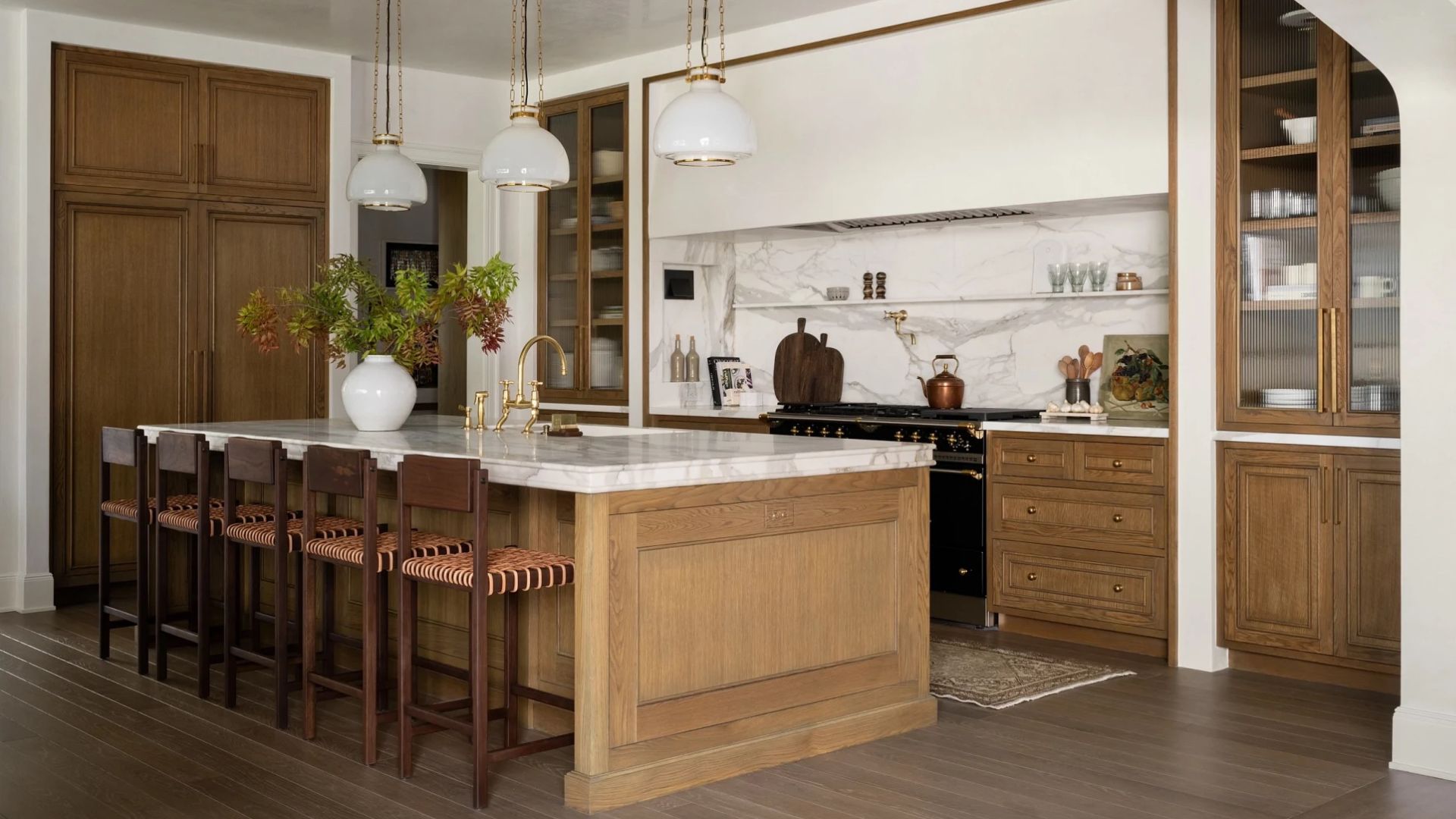 A white kitchen with a marble backsplash and floating shelves, wood cupboards with glass cabinets, three pendant lamps over a wood island with a marble top and woven wood chairs lined up by the side