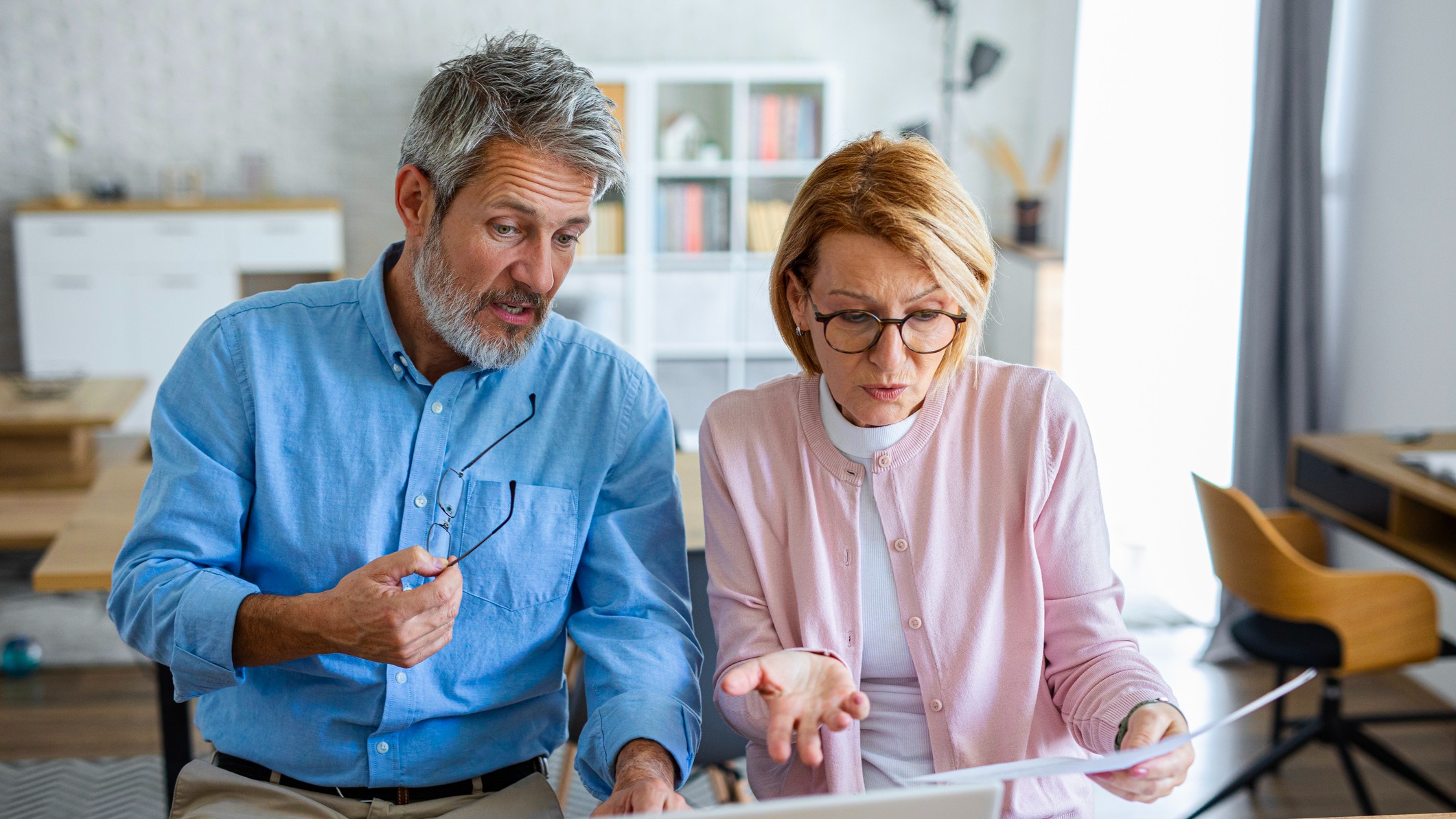 An older couple sits in front of a laptop surrounded by documents, visibly pressured as they attempt to organize their finances or retirement plan.