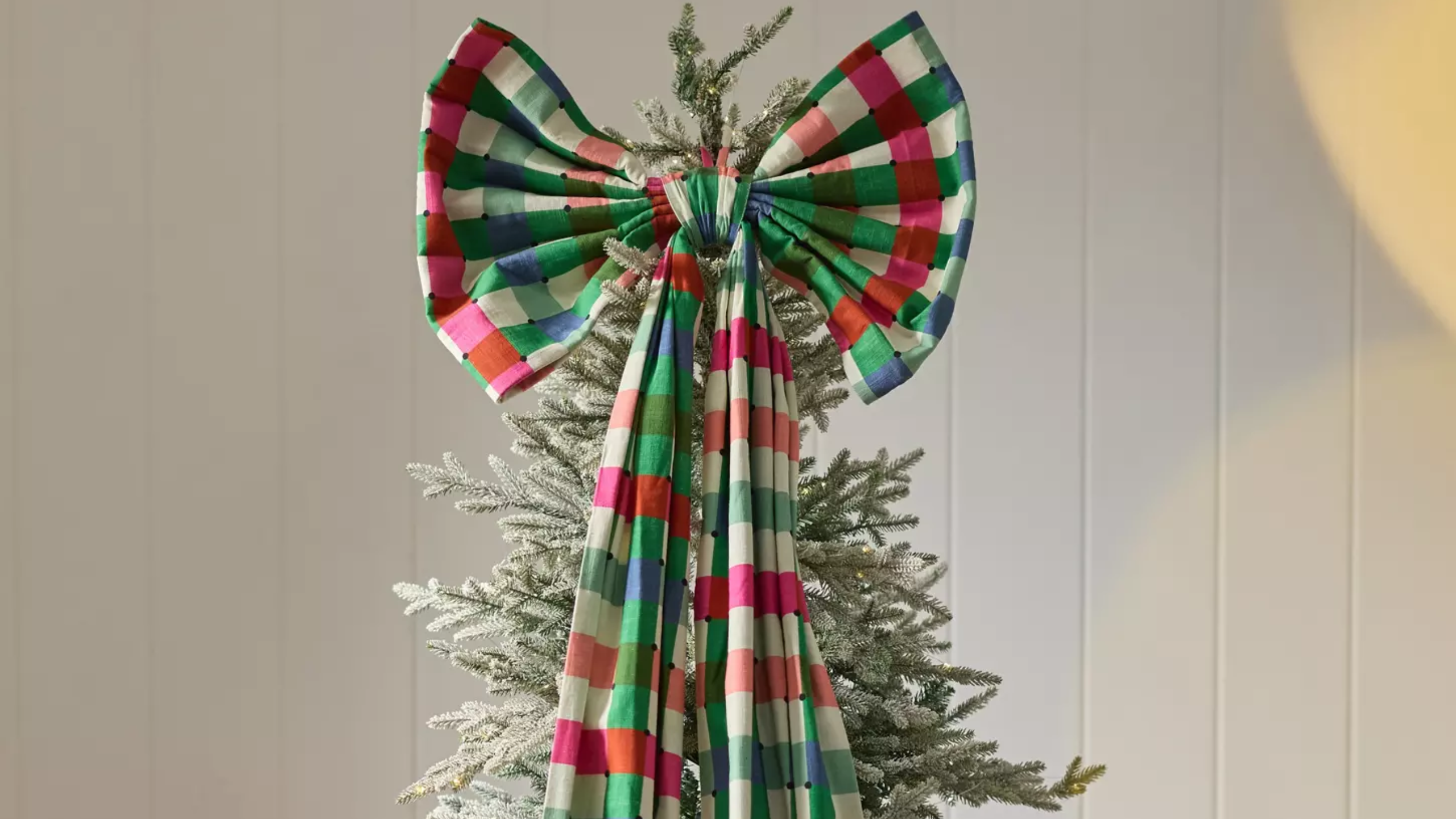 Anthropologie giant plaid bow tree topper crowning the top of a faux frosted Christmas tree, grounded by a wool-and-cotton-blend patchwork plaid skirt.