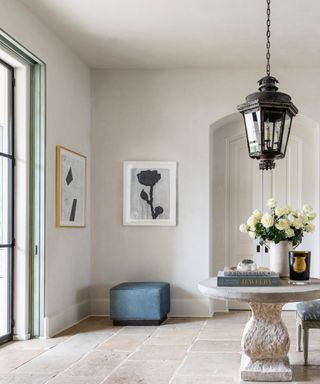 hallway with stone floor tiles, stone circular table with flowers and books, stool, vintage lantern, dresser with mirror, flowers, artwork, crittall doors to left,