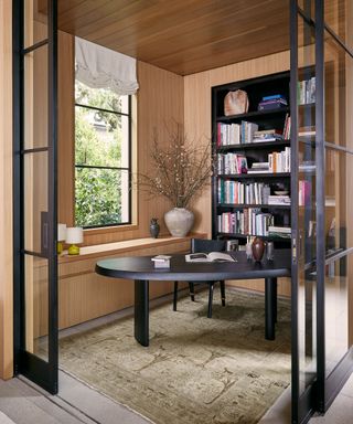 An office space with wooden walls and a black bookshelf, table and chair in front of a window with glass sliding doors that meet on a corner