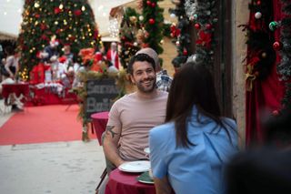 Jack (Shayne Ward) and Kate (Catherine Tyldesley) are sitting at a table outside a cafe. Kate has her back to the camera, Jack looks cheerful. In the background is an enormous, lavishly-decorated Christmas tree with a grotto in front of it, and a sandwich board with a message in Croatian.