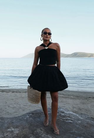 A woman wearing black sunglasses, large hoops, a black halter top, a black balloon skirt, gold sandals, and a woven tote bag.