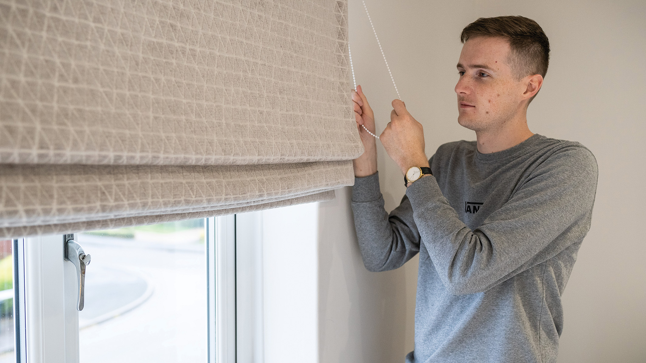 A man in a gray sweater adjusts beige Roman blinds by a window. He looks focused, gently pulling the cord. Soft natural light filters in, creating a calm atmosphere