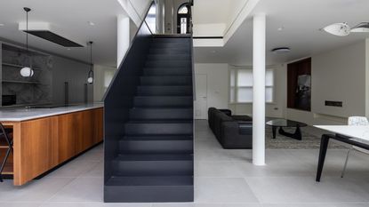 A minimalist, open plan kitchen with a wooden island, stone tiles, and a black staircases in the center
