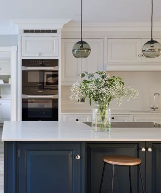 A kitchen with a kitchen island with a white counter and a navy blue back panel, a glass vase with green stems, and white cabinets and a black integrated oven behind this