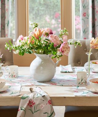 A wooden dining table with pink and green flower patterned fabrics and placemats with a white ceramic bowl of pink flowers, with a window with curtains in the background