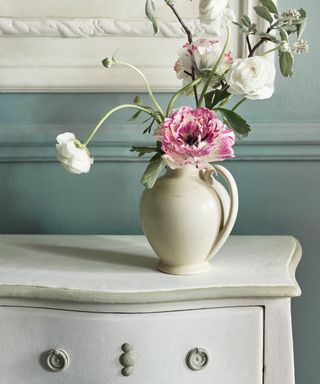 A white drawer set with a white vase with flowers, in front of a blue wall
