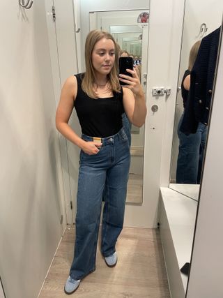 Woman in dressing room wears black top, blue jeans and blue trainers