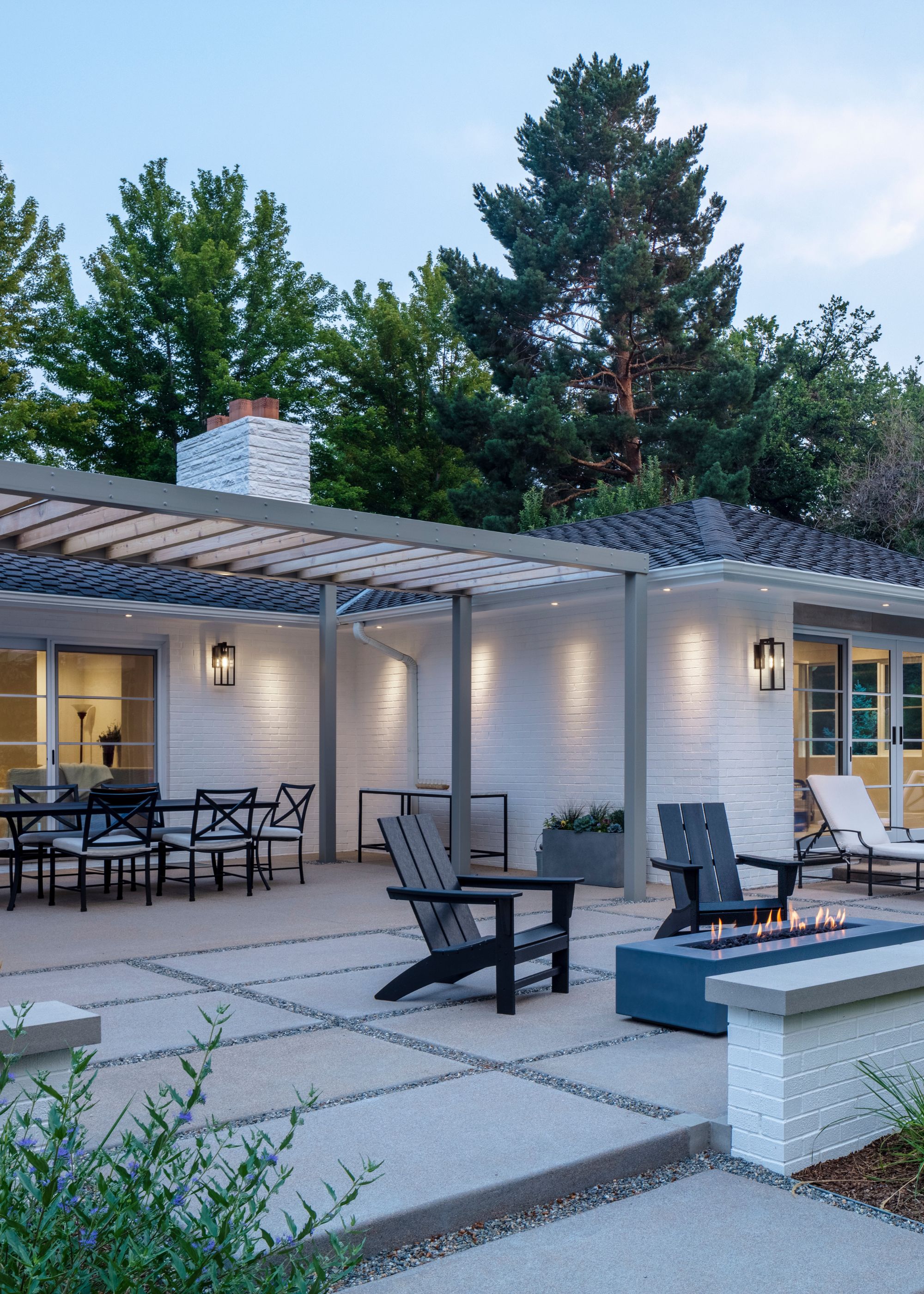 A patio with a small fire pit and two arm chairs place in front of it. There is also a view of a the pergola that also have a dining table and chairs beneath it.
