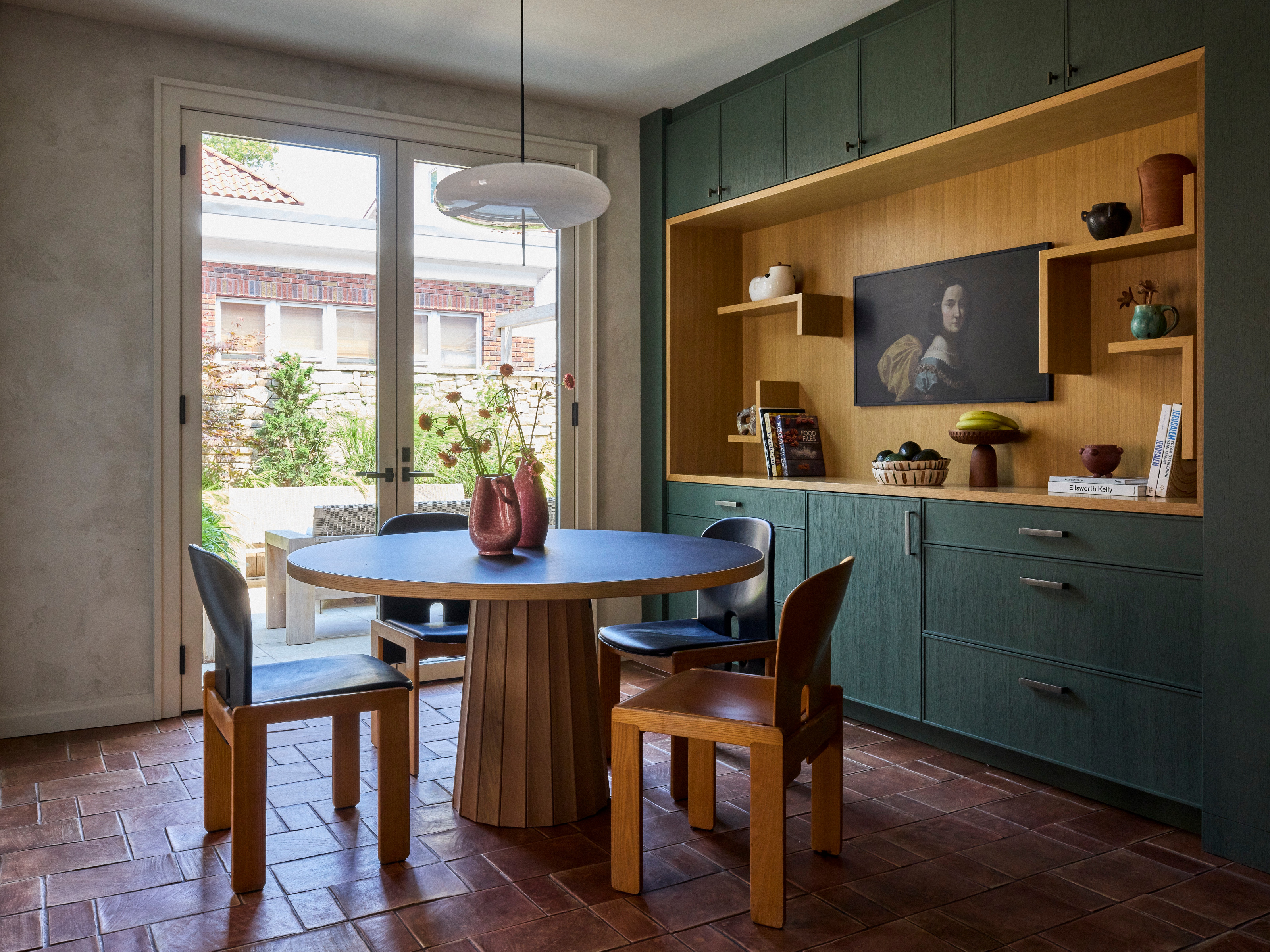 Dining area with teal stained wood kitchen units, terracotta tiled floor, round pedestal dining table and chunky wood chairs