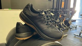 A pair of black GoRuck ballistic trainers in a gym; behind them we see assault bikes and a white board.