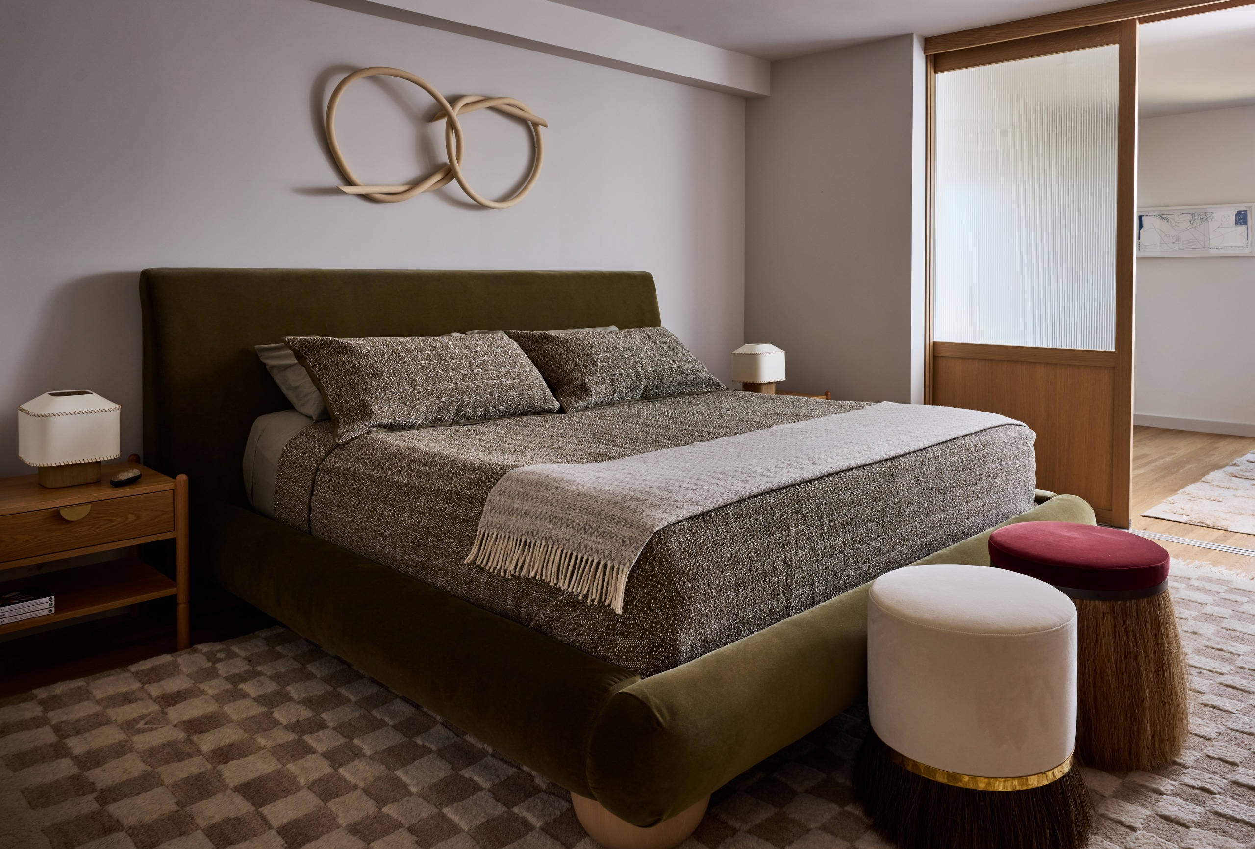 A bedroom with a khaki velvet bed, two pouffes at the foot of the bed and a neutral chequered rug underneath. A sliding wood and glass door to go through to the next room