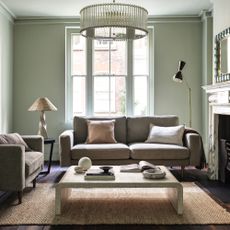 how to decorate a north-facing room, pale green living room with taupe matching sofas, dark stained wooden floorboards, jute rug, curved coffee table, retro chandelier pendant, rattan table lamp