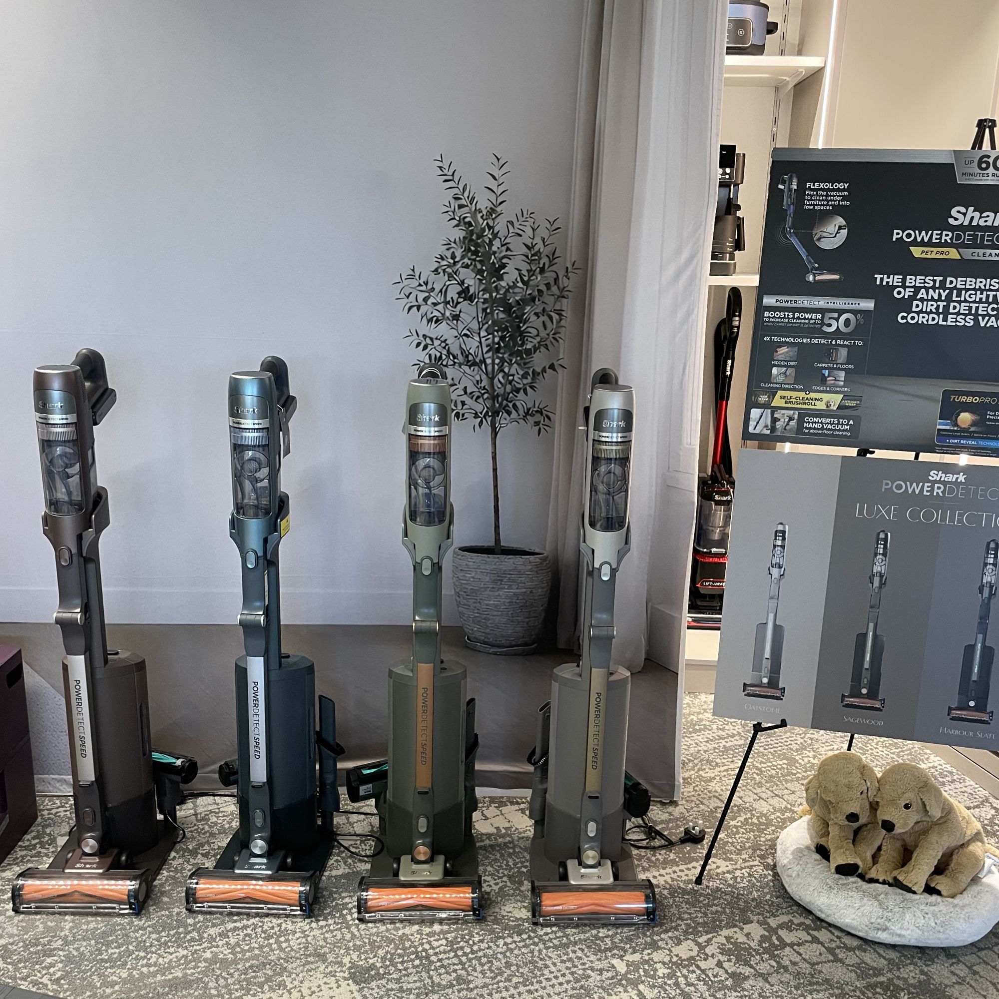 Four vacuums from the Shark PowerDetect Speed Luxe Collection sitting on carpet, next to a board of information about the vacuums